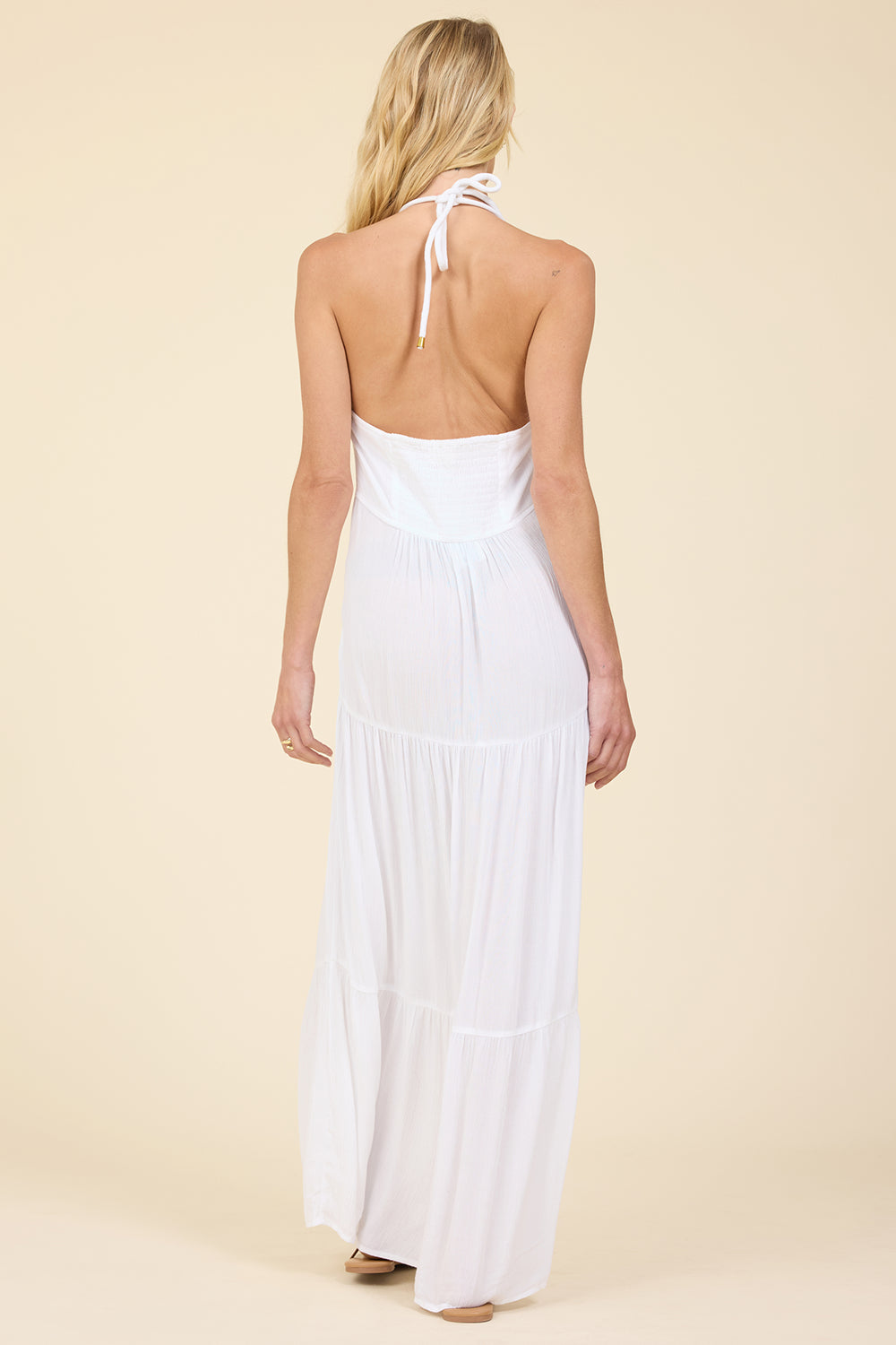 A woman viewed from the back wearing a white halter-neck maxi dress with an open back and tie straps, standing against a plain beige studio background.