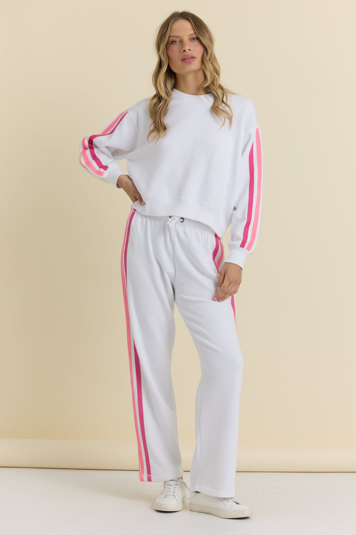 A woman modeling a white two-piece tracksuit with bright neon pink stripes down the sleeves and legs, paired with white sneakers, against a solid beige studio background.