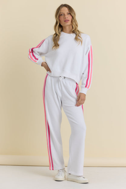 A woman modeling a white two-piece tracksuit with bright neon pink stripes down the sleeves and legs, paired with white sneakers, against a solid beige studio background.