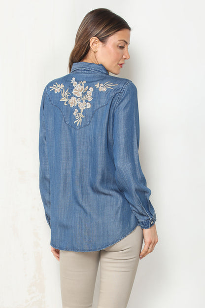 A woman shown from the back wearing a blue dark wash denim shirt with white floral embroidery on the upper back, paired with beige pants, against a plain white background.