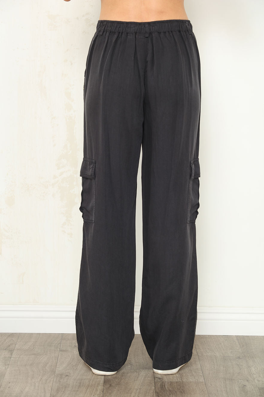 Black wide-leg cargo pants with side flap pockets, shown on a model back view from the waist down against a light beige wall and wood floor.