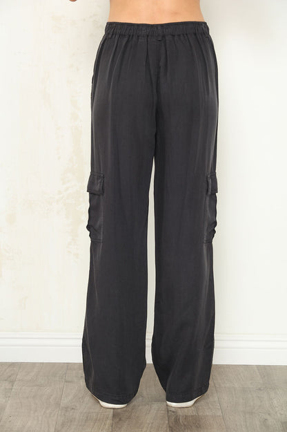 Black wide-leg cargo pants with side flap pockets, shown on a model back view from the waist down against a light beige wall and wood floor.