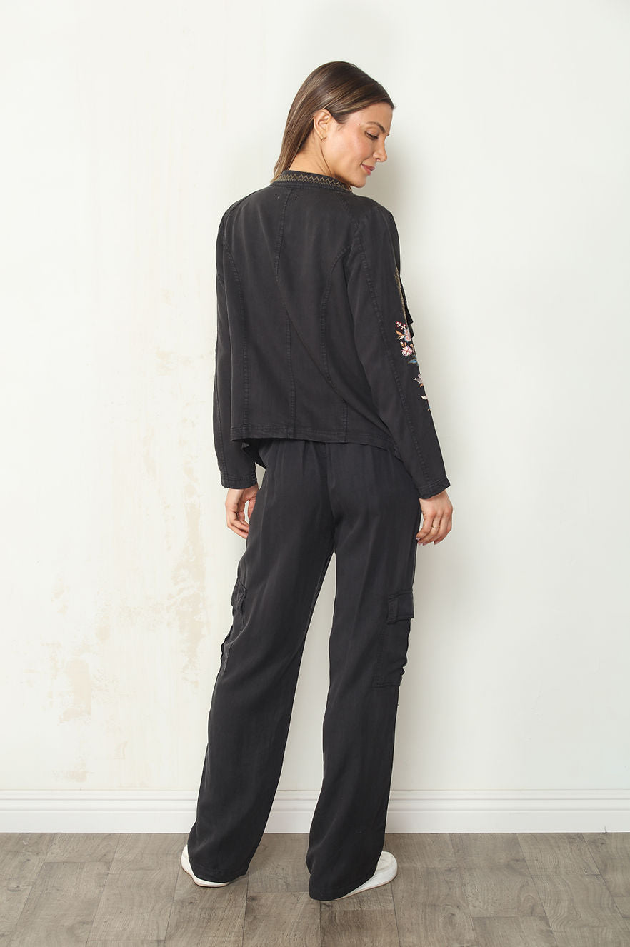 A woman stands with her back to the camera wearing a black long-sleeve cropped jacket with floral embroidery on the sleeve and matching black wide-leg cargo pants, set against a plain off-white wall and light wood floor.