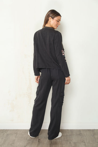 A woman stands with her back to the camera wearing a black long-sleeve cropped jacket with floral embroidery on the sleeve and matching black wide-leg cargo pants, set against a plain off-white wall and light wood floor.