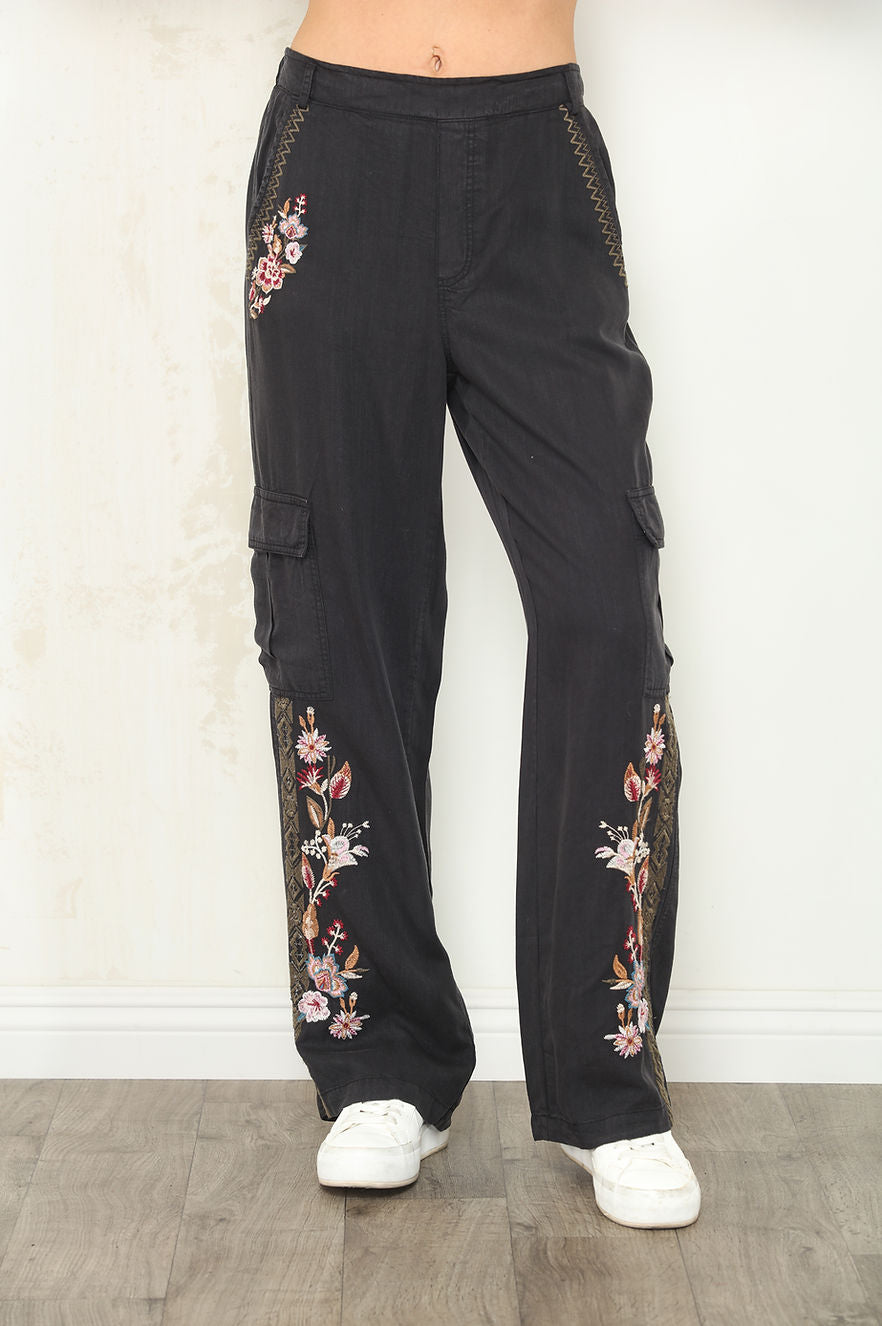 Front view of Black wide-leg cargo pants with floral embroidery on the legs and side pockets, shown on a model wearing white sneakers against a light wall and wood floor.