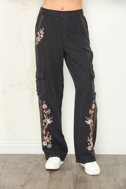 Front view of Black wide-leg cargo pants with floral embroidery on the legs and side pockets, shown on a model wearing white sneakers against a light wall and wood floor.