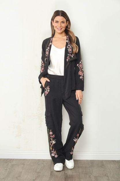 A woman modeling a black two-piece lounge set with pink floral embroidery on the sleeves and legs, featuring a zip-up jacket and matching cargo pants, paired with a white tank top and white sneakers.