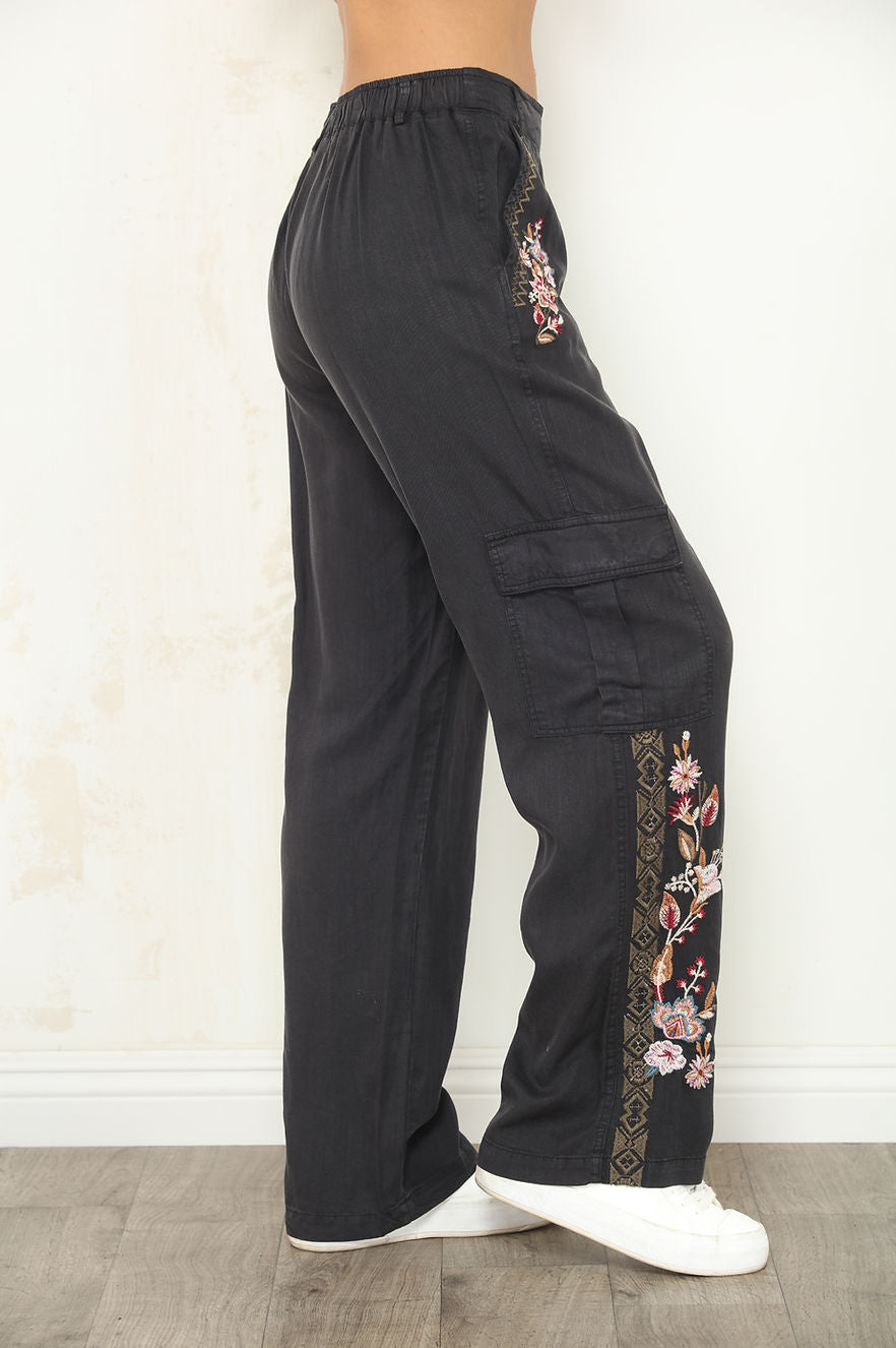 Black wide-leg cargo pants with floral embroidery on the side pockets and lower hems, shown on a model from the waist down wearing white sneakers.