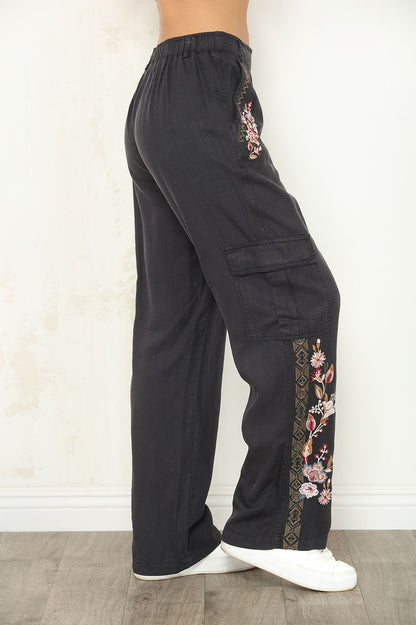 Black wide-leg cargo pants with floral embroidery on the side pockets and lower hems, shown on a model from the waist down wearing white sneakers.