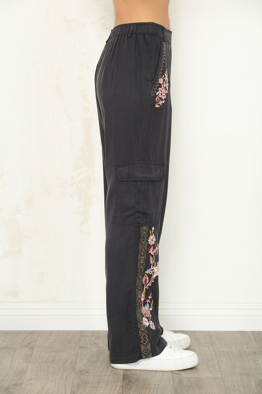 Black wide-leg cargo pants with floral embroidered details on the sides, shown on a model from the waist down wearing white sneakers against a light textured wall.