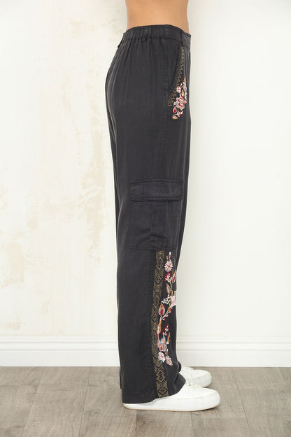 Black wide-leg cargo pants with floral embroidered details on the sides, shown on a model from the waist down wearing white sneakers against a light textured wall.