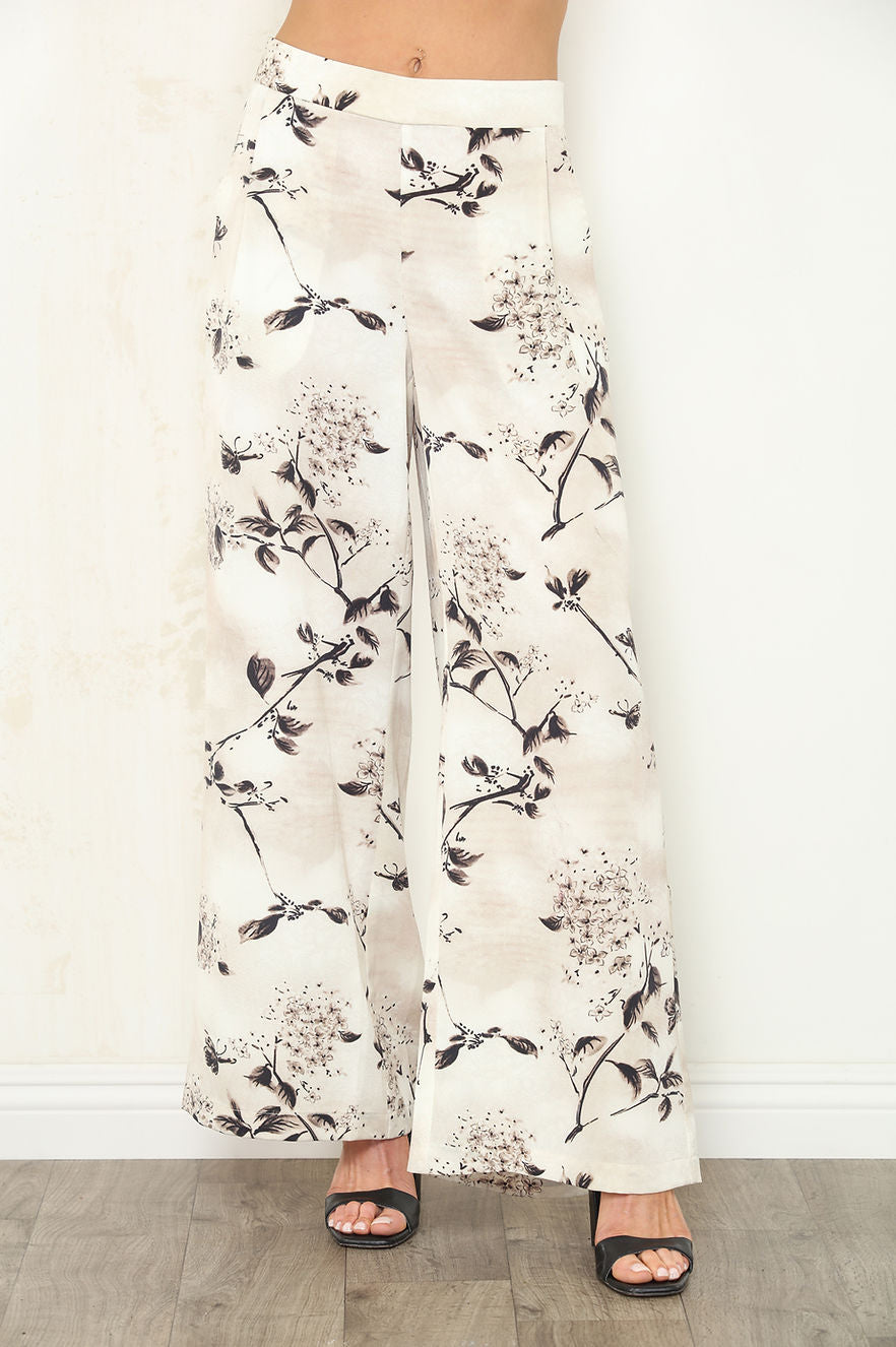 A person wearing wide-leg pants with a black floral and branch Faded Petal print on an off-white background, paired with black slide sandals, standing against a white wall.