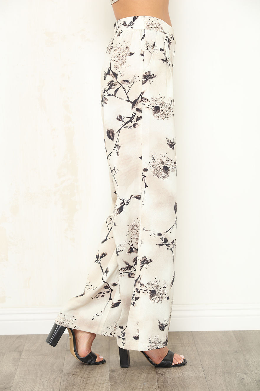 A person shown from the waist down wearing high-waisted, wide-leg pants with a black floral Faded Petal print on an off-white background, paired with black heeled sandals.