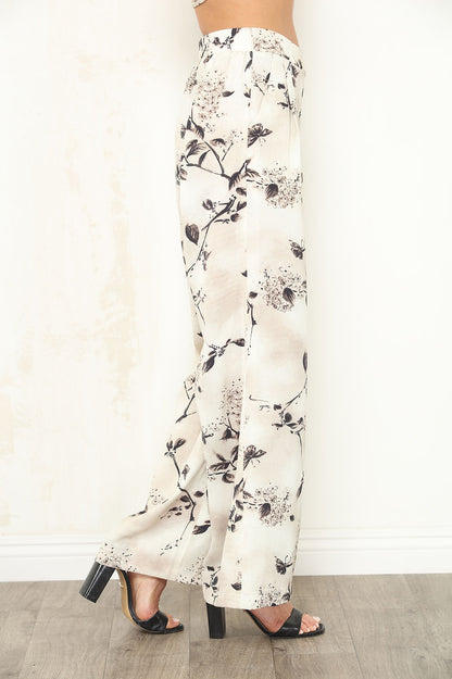 A person shown from the waist down wearing high-waisted, wide-leg pants with a black floral Faded Petal print on an off-white background, paired with black heeled sandals.
