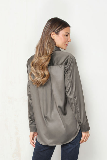 Back view of a woman modeling the camelia long-sleeve button-up shirt with a dark olive green base and a large, vibrant floral print in red, beige, and gray, paired with dark blue jeans against a plain white wall.