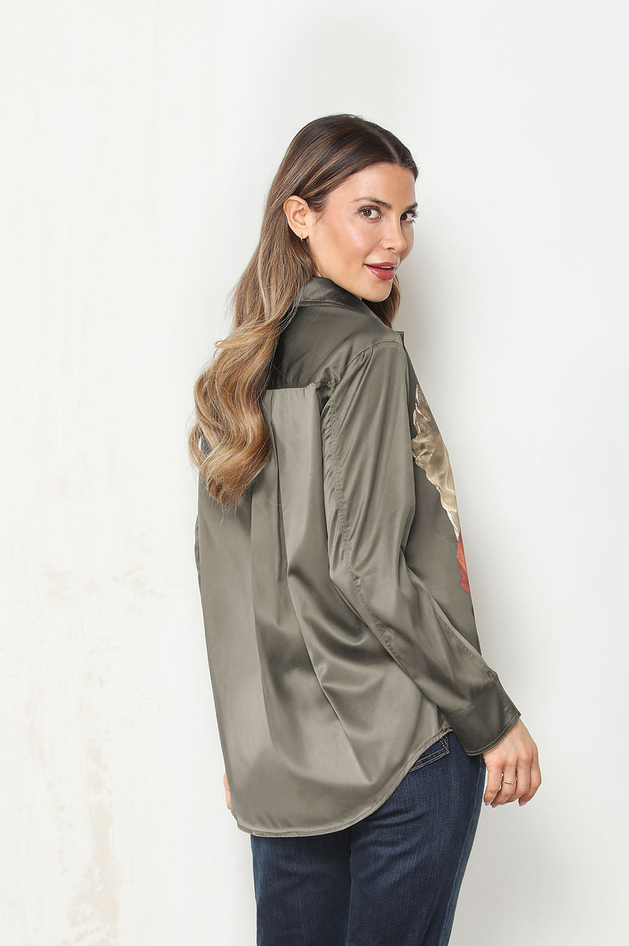 Back view of a woman modeling the camelia long-sleeve button-up shirt with a dark olive green base and a large, vibrant floral print in red, beige, and gray, paired with dark blue jeans against a plain white wall.