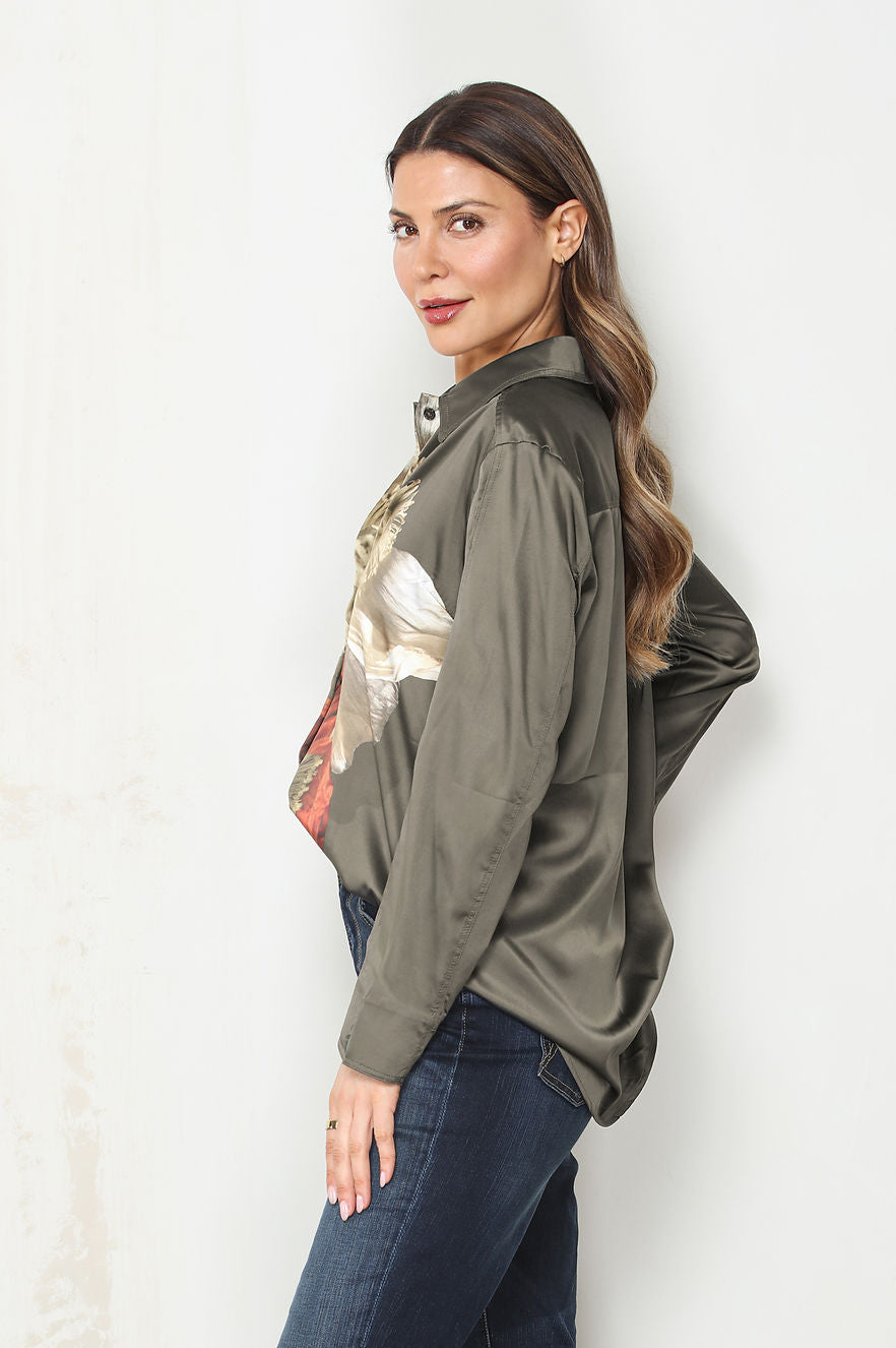 Side view of a woman modeling the camelia long-sleeve button-up shirt with a dark olive green base and a large, vibrant floral print in red, beige, and gray, paired with dark blue jeans against a plain white wall.