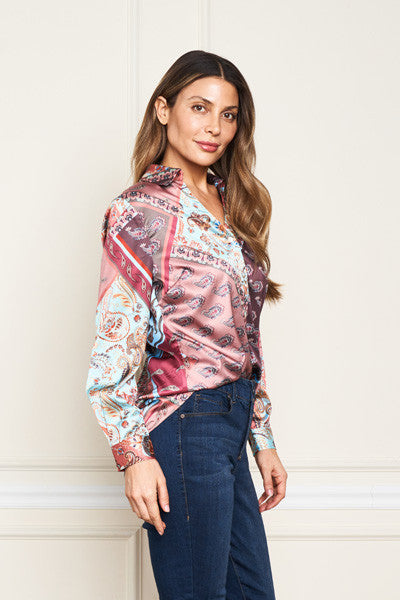 A woman modeling a long-sleeve button-up shirt with a multicolor paisley and bandana print in pink, blue, and white, paired with dark blue jeans against a cream paneled wall.