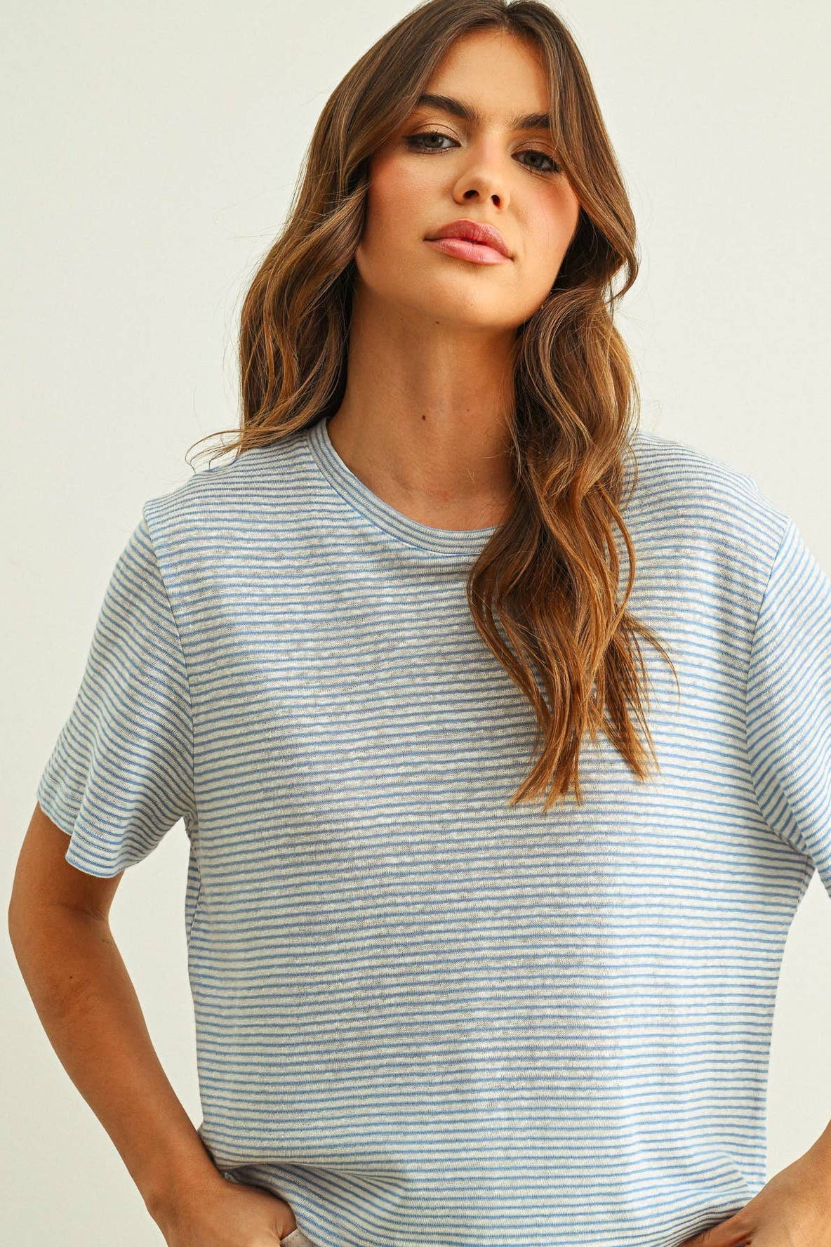 A woman wearing a light blue and white horizontally striped short-sleeve slub t-shirt, photographed against a plain off-white background.