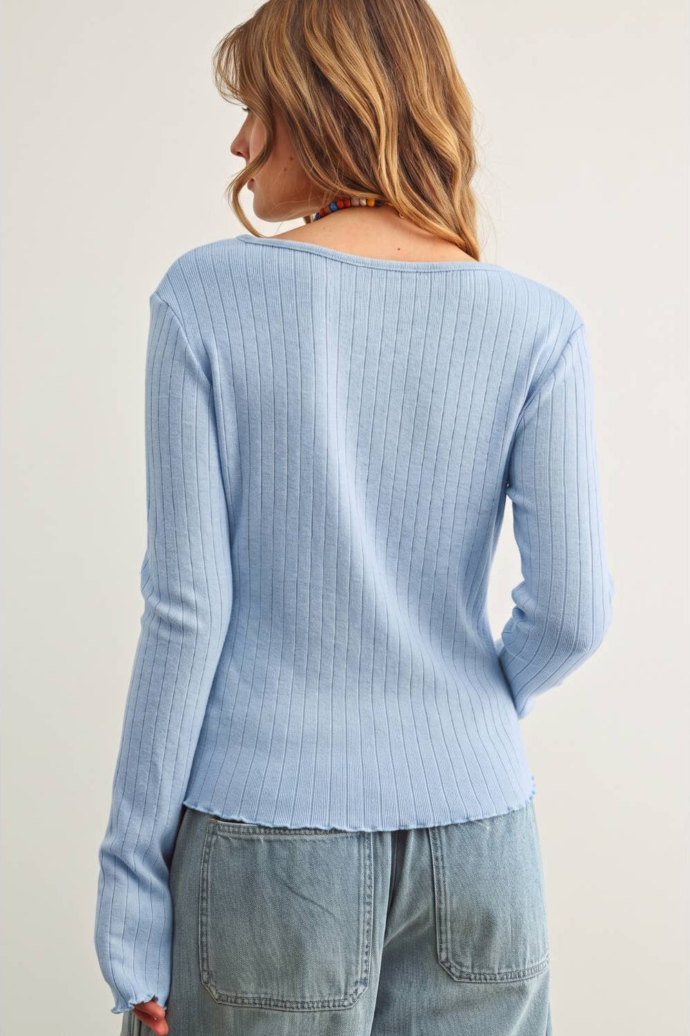 A woman shown from the back wearing a light blue ribbed knit sweater and light-wash jeans against a plain light beige studio background.