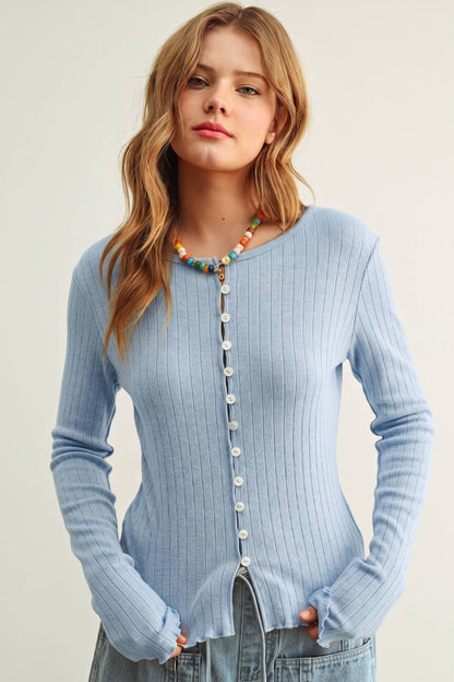 A woman wearing a light blue ribbed long-sleeve cardigan with a button-down front, styled with a colorful beaded necklace and light-wash jeans against a plain light beige background.