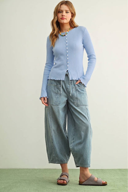 A woman modeling a light blue ribbed knit cardigan with button details, paired with wide-leg light blue denim pants and brown slide sandals, set against a neutral studio background.