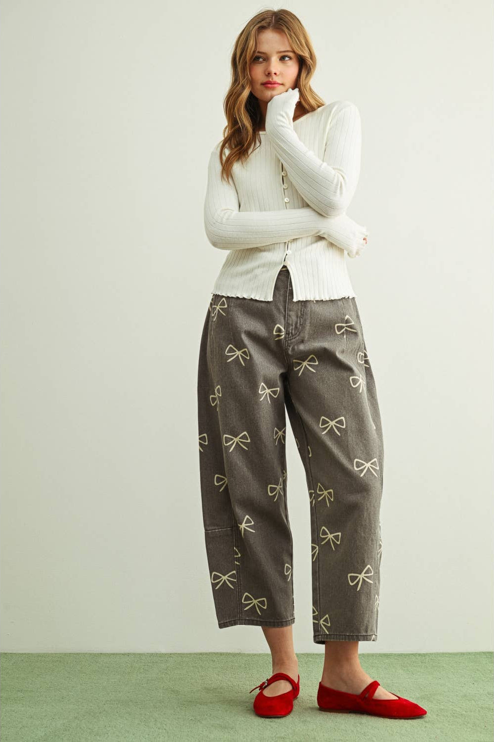 A woman stands against a plain light wall wearing a cream ribbed long-sleeve top, wide-leg cropped pants in dark gray with a repeating bow print, and red flat shoes.