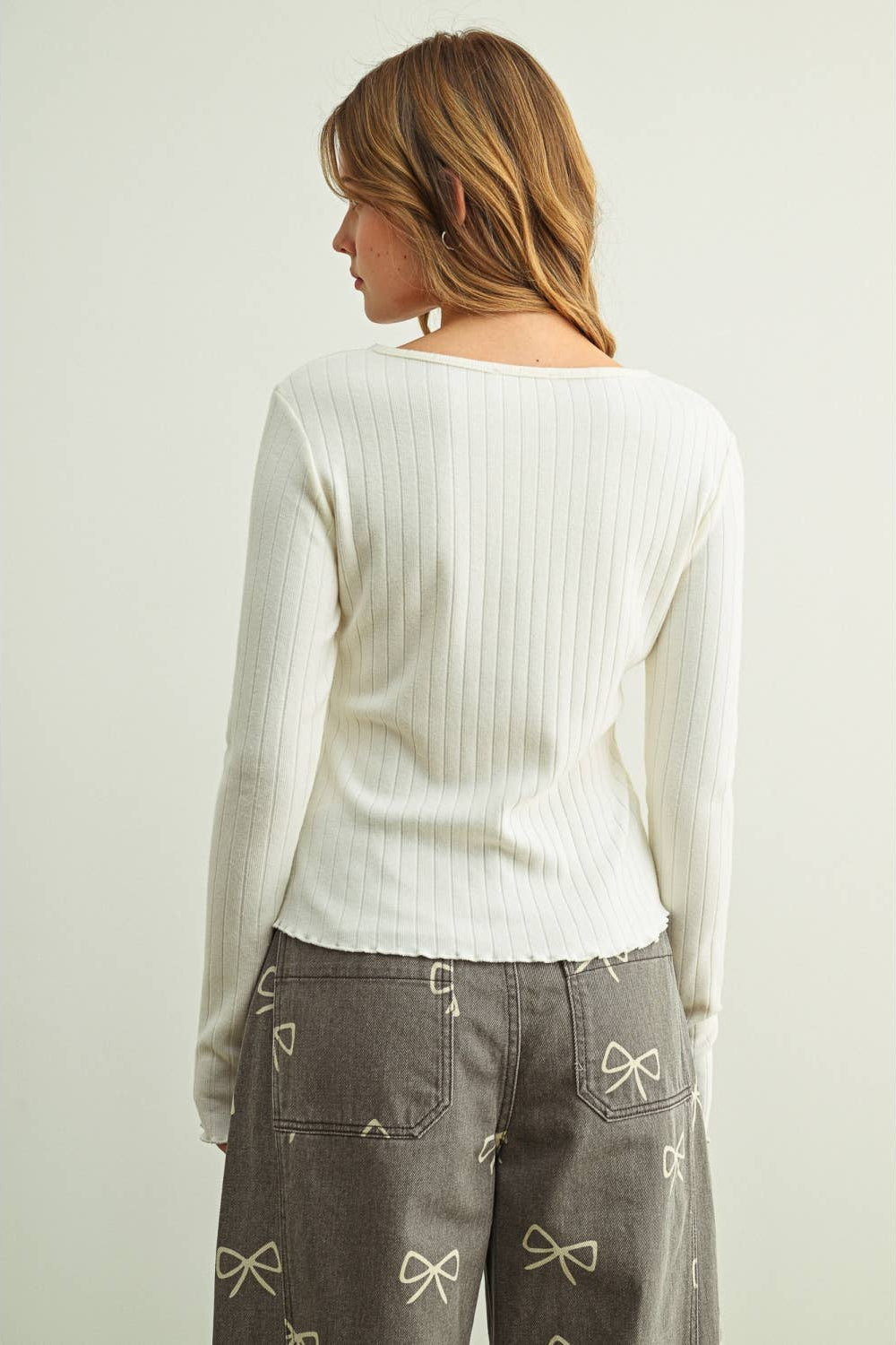 A woman shown from the back wearing a cream ribbed long-sleeve top and gray pants with bow embroidery, against a plain light background.
