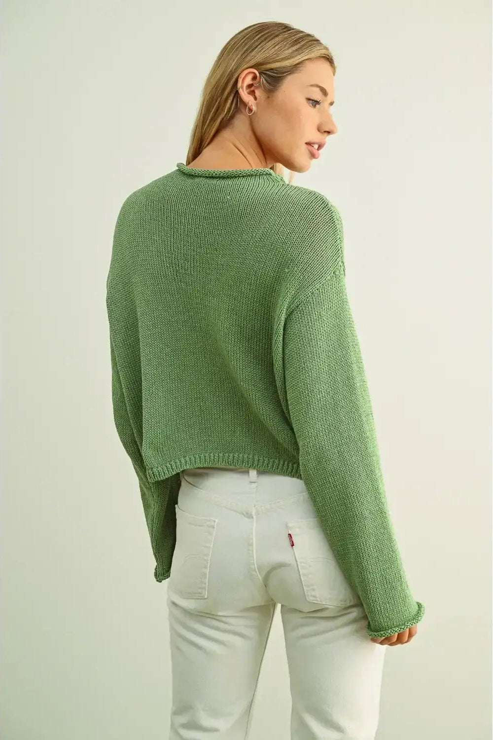 Back view of a woman wearing a matcha green cropped knit sweater and white pants, shown from a back-three-quarter angle against a plain light gray studio background.