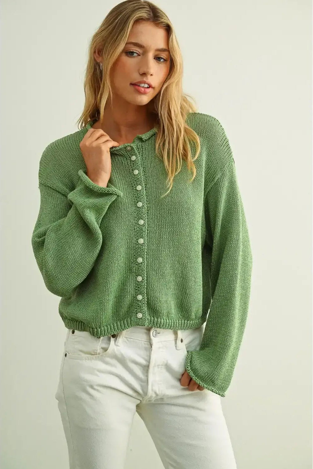 A woman wearing a matcha green knit button-up cardigan sweater and white jeans, standing against a light beige studio background.