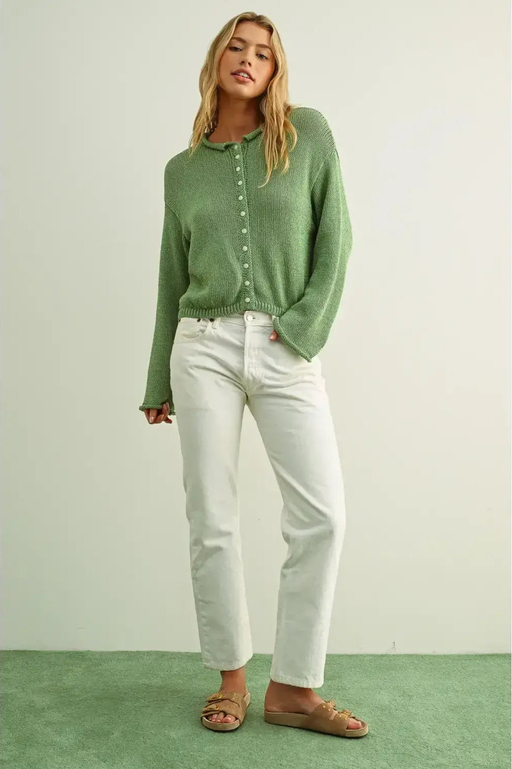 A woman modeling a matcha green knit button-front cardigan sweater with white cropped jeans and tan flat sandals, standing against a light neutral wall with a green floor.