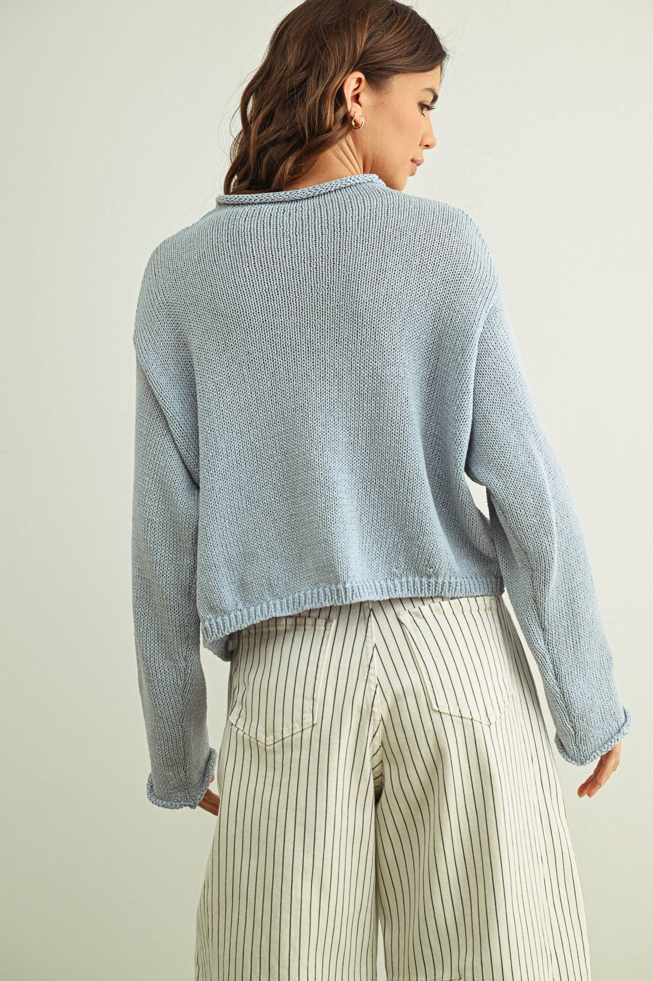 Back view of woman wearing a sky blue sweater and white pants with black stripes on a plain background