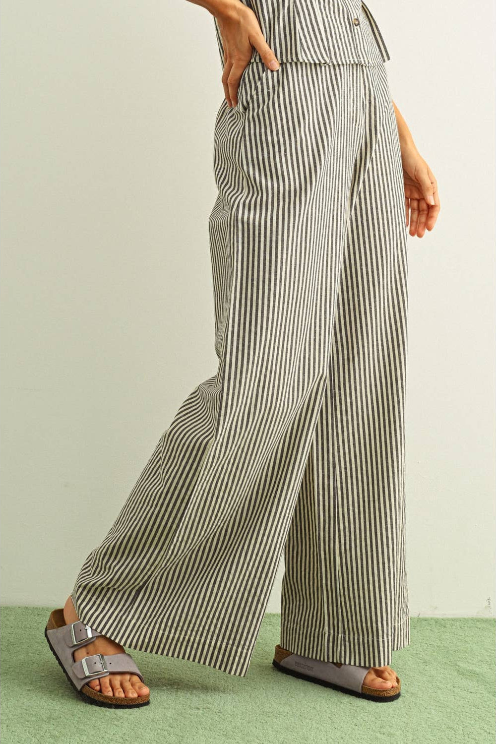 A person wearing high-waisted, wide-leg trousers with vertical black and beige stripes, paired with brown slide sandals, shown from the waist down against a light neutral wall and green textured floor.