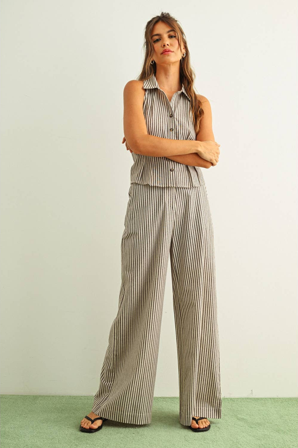 A woman modeling a sleeveless button-front top and matching wide-leg pants set with thin vertical brown and white stripes, standing against a plain light wall on a green floor.