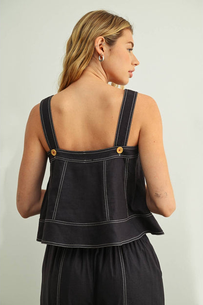 A woman shown from the back wearing a navy top with wide shoulder straps, gold-tone buttons, and contrasting white stitching, against a plain light gray studio background.