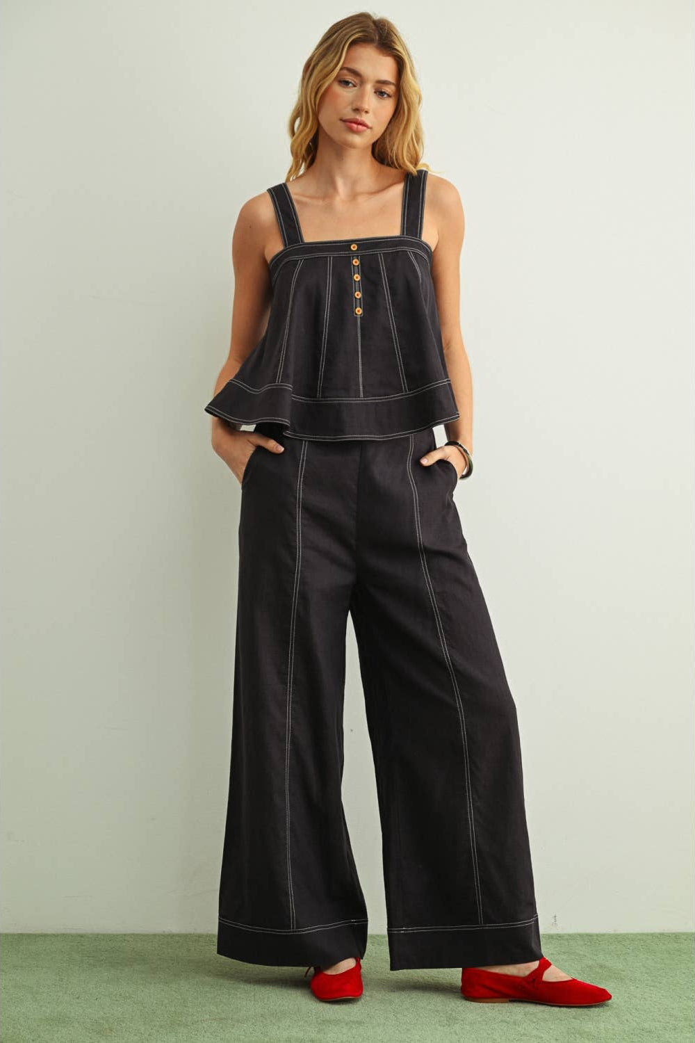 A woman modeling a navy sleeveless top and matching wide-leg pants with contrast white stitching and gold-tone buttons, paired with red flats, against a light gray wall and green floor.