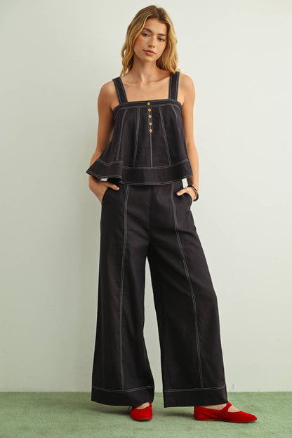 A woman modeling a navy sleeveless top and matching wide-leg pants with contrast white stitching and gold-tone buttons, paired with red flats, against a light gray wall and green floor.