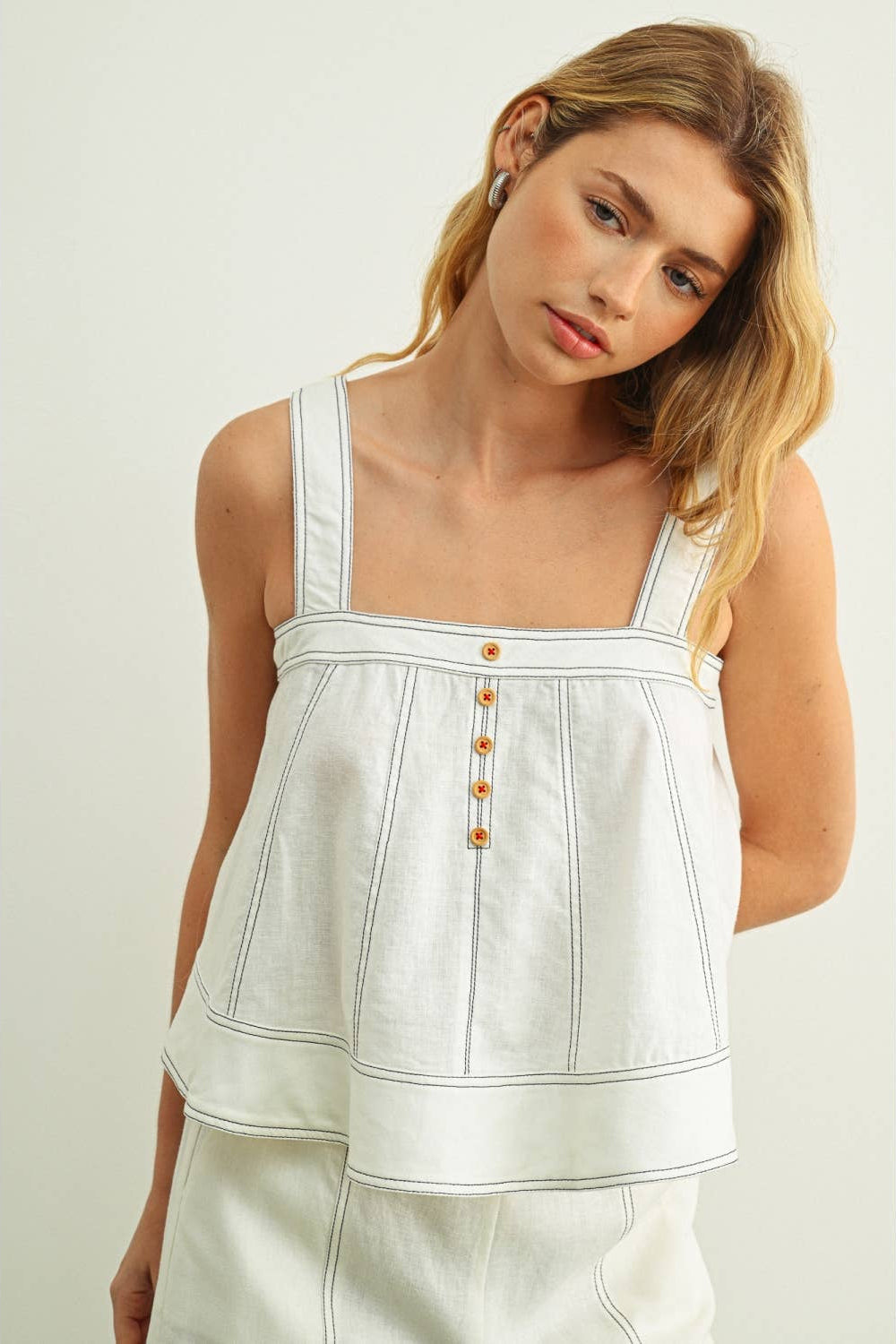 Woman wearing a white sleeveless top with stitching & button details on a light background