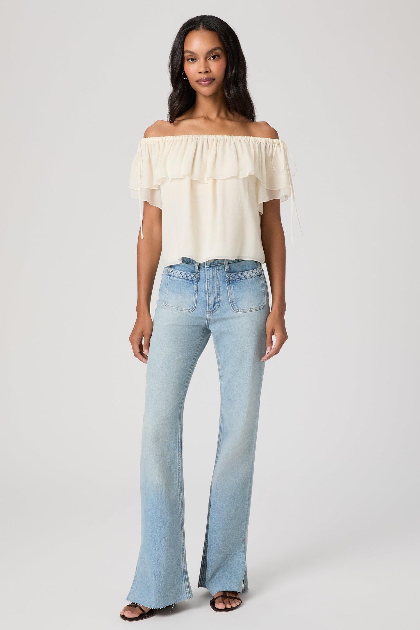 Woman wearing a white off-shoulder top and light blue montebello sofia jeans on a white background