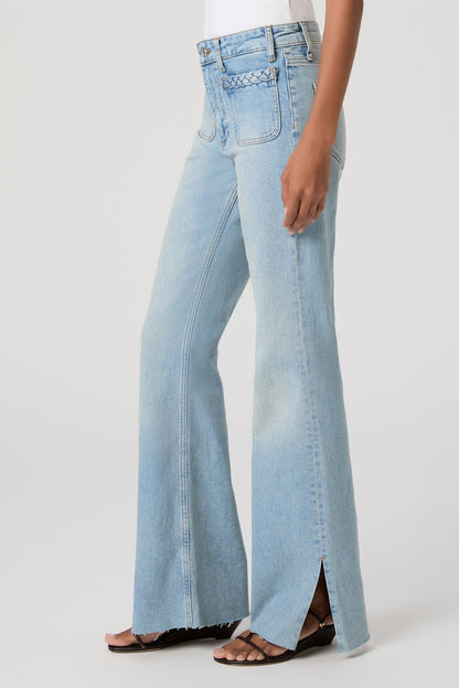 A side view of a model wearing light-wash montebello blue sofia flare jeans with a braided waistband detail and front slit details, paired with brown slide sandals against a plain light gray studio background.