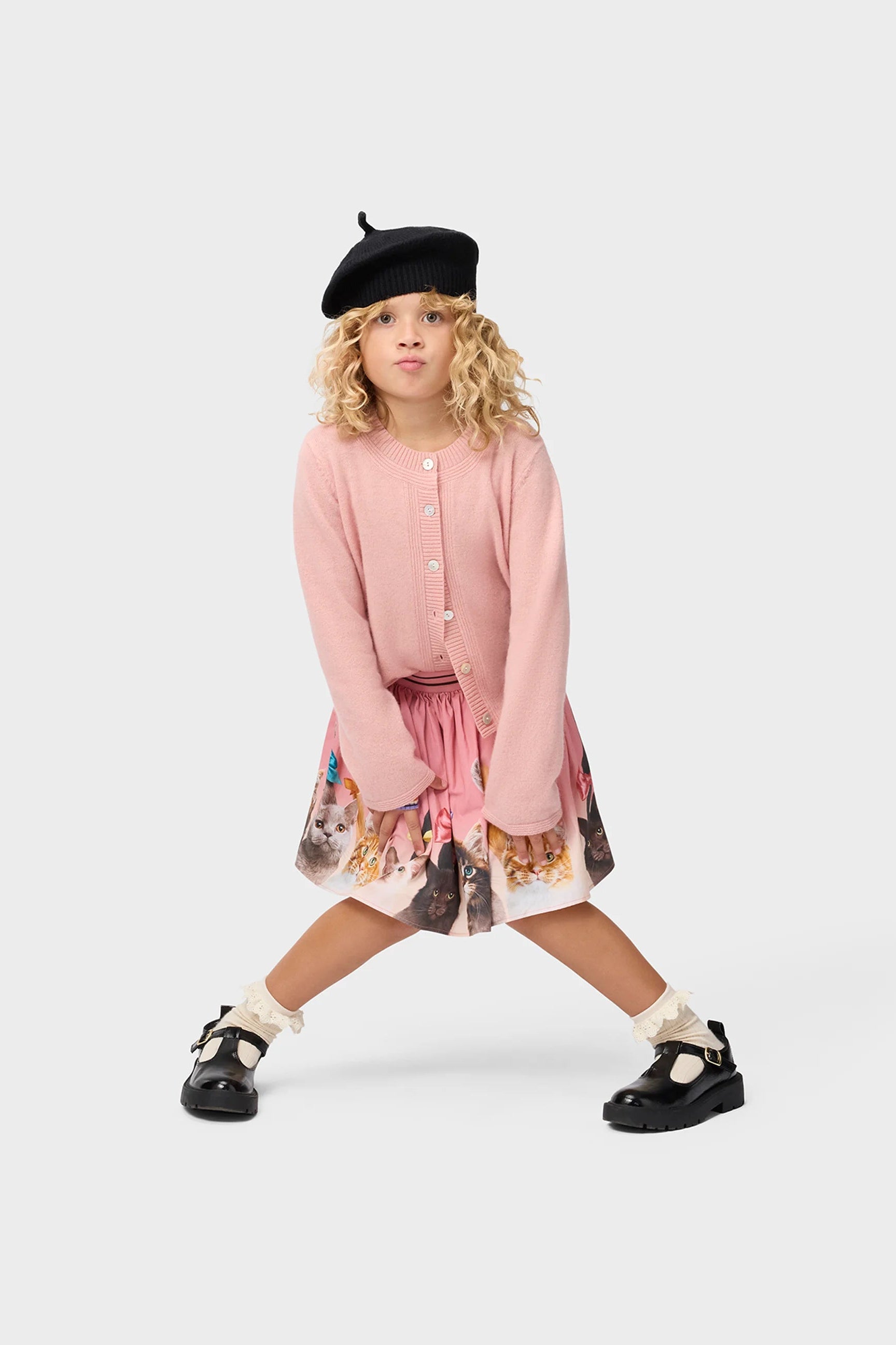 A young girl modeling a pink cardigan and patterned skirt set, styled with a black beret, white socks, and black Mary Jane shoes, posed against a light gray studio background.