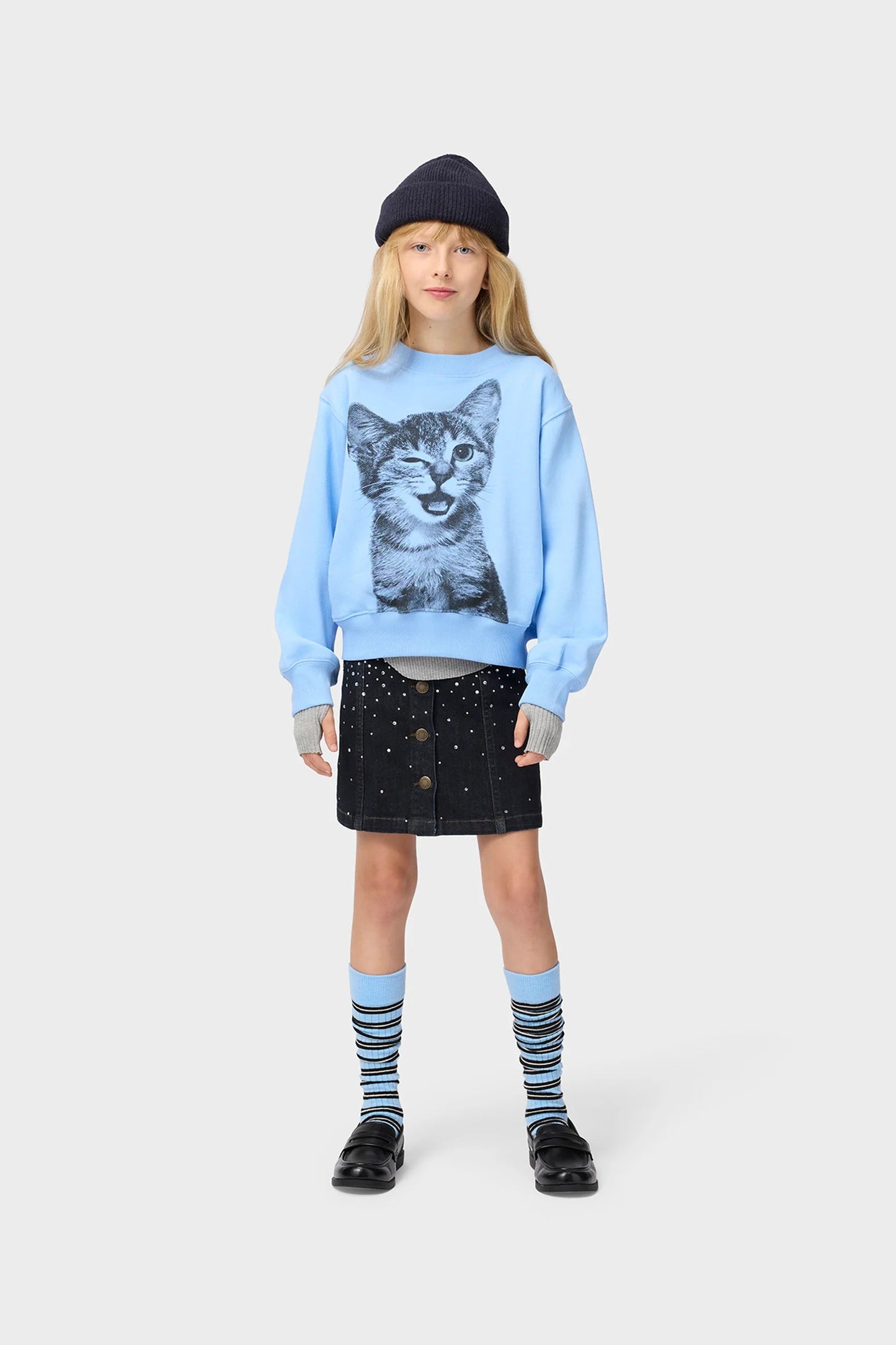 A young girl stands against a light gray studio background wearing a light blue sweatshirt with a large black-and-white cat face graphic on the front, a dark beanie, a dark skirt, striped socks, and black shoes.