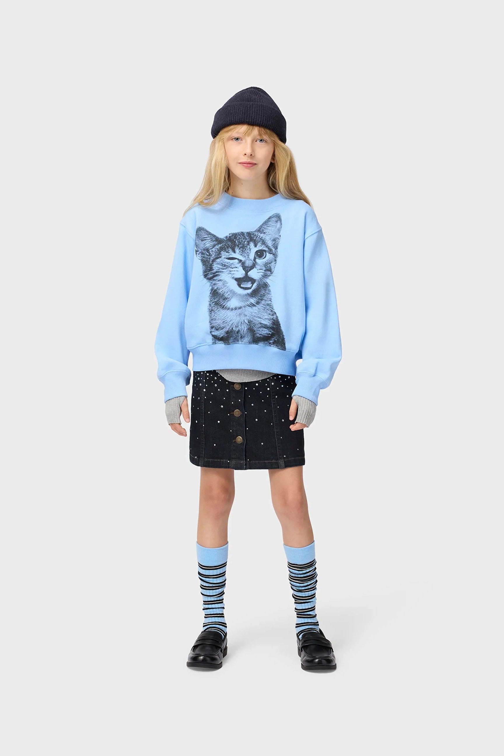 A young girl stands against a light gray studio background wearing a light blue sweatshirt with a large black-and-white cat face graphic on the front, a dark beanie, a dark skirt, striped socks, and black shoes.