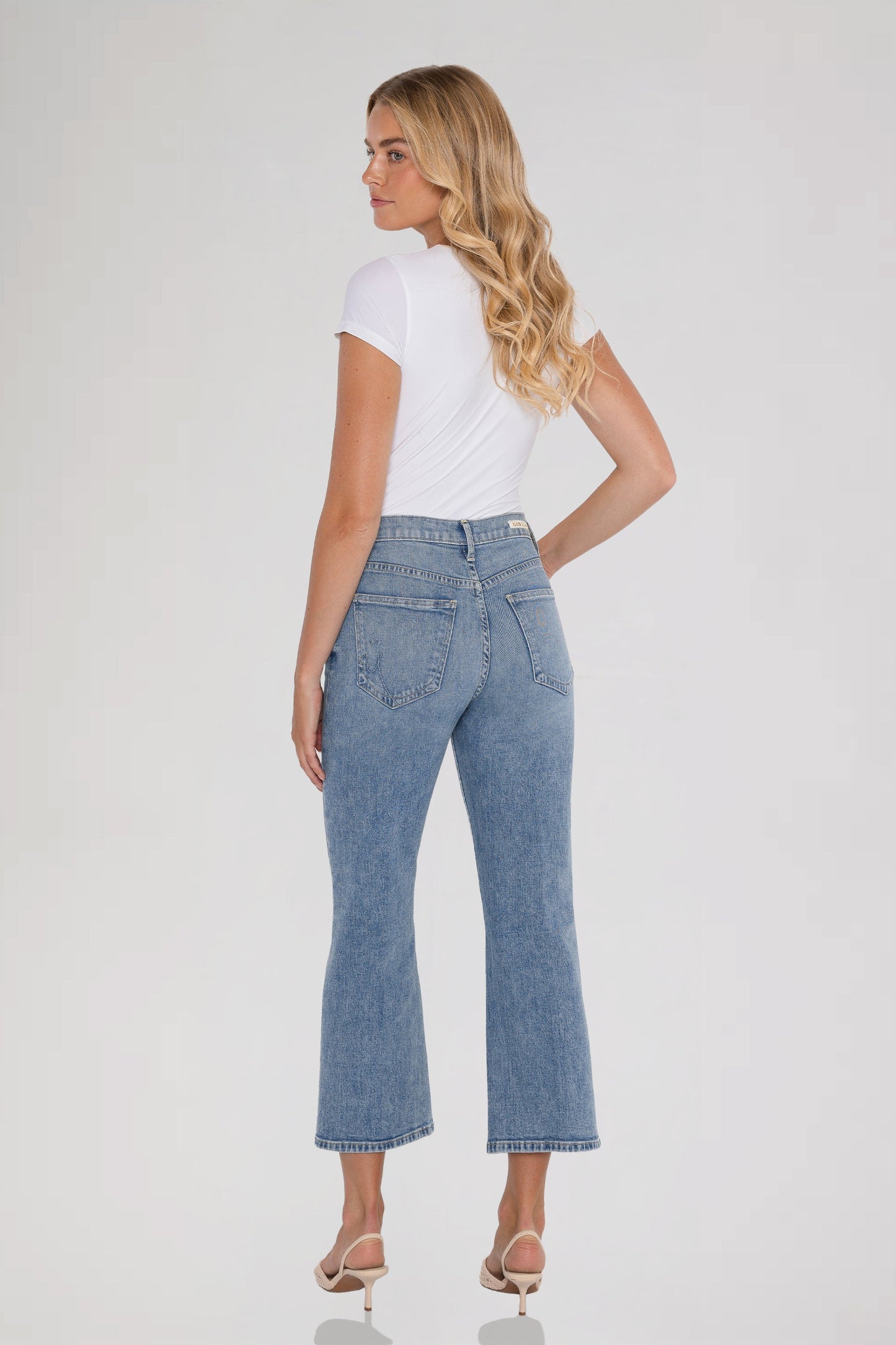 A woman is shown from the back wearing light-wash bleu ciel cropped flare jeans, a white short-sleeve top, and beige high-heeled sandals against a plain light gray studio background.