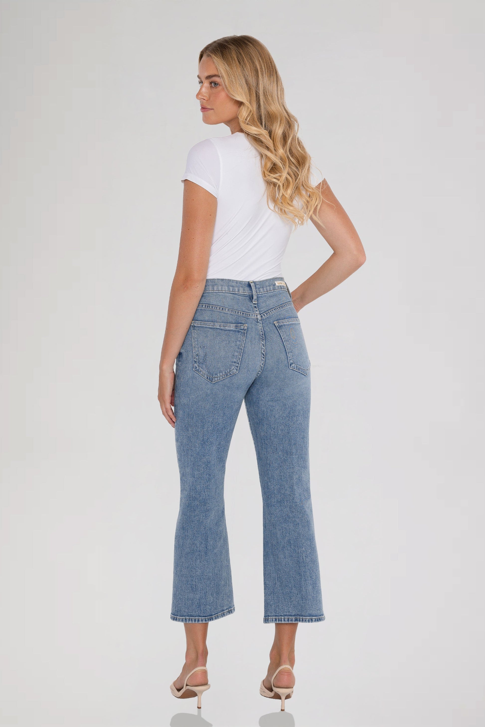 A woman is shown from the back wearing light-wash bleu ciel cropped flare jeans, a white short-sleeve top, and beige high-heeled sandals against a plain light gray studio background.