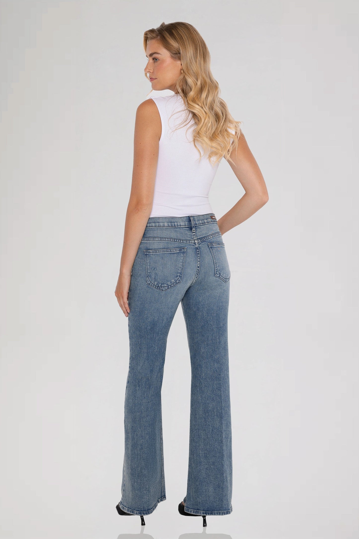 A woman is shown from the back wearing  a white sleeveless top and bleu perle grace mid rise jeans standing against a plain background