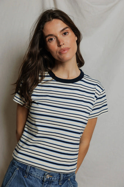 A woman wearing a navy and white pacific  striped t-shirt with a dark navy collar, paired with blue jeans, posing against a draped white fabric backdrop.