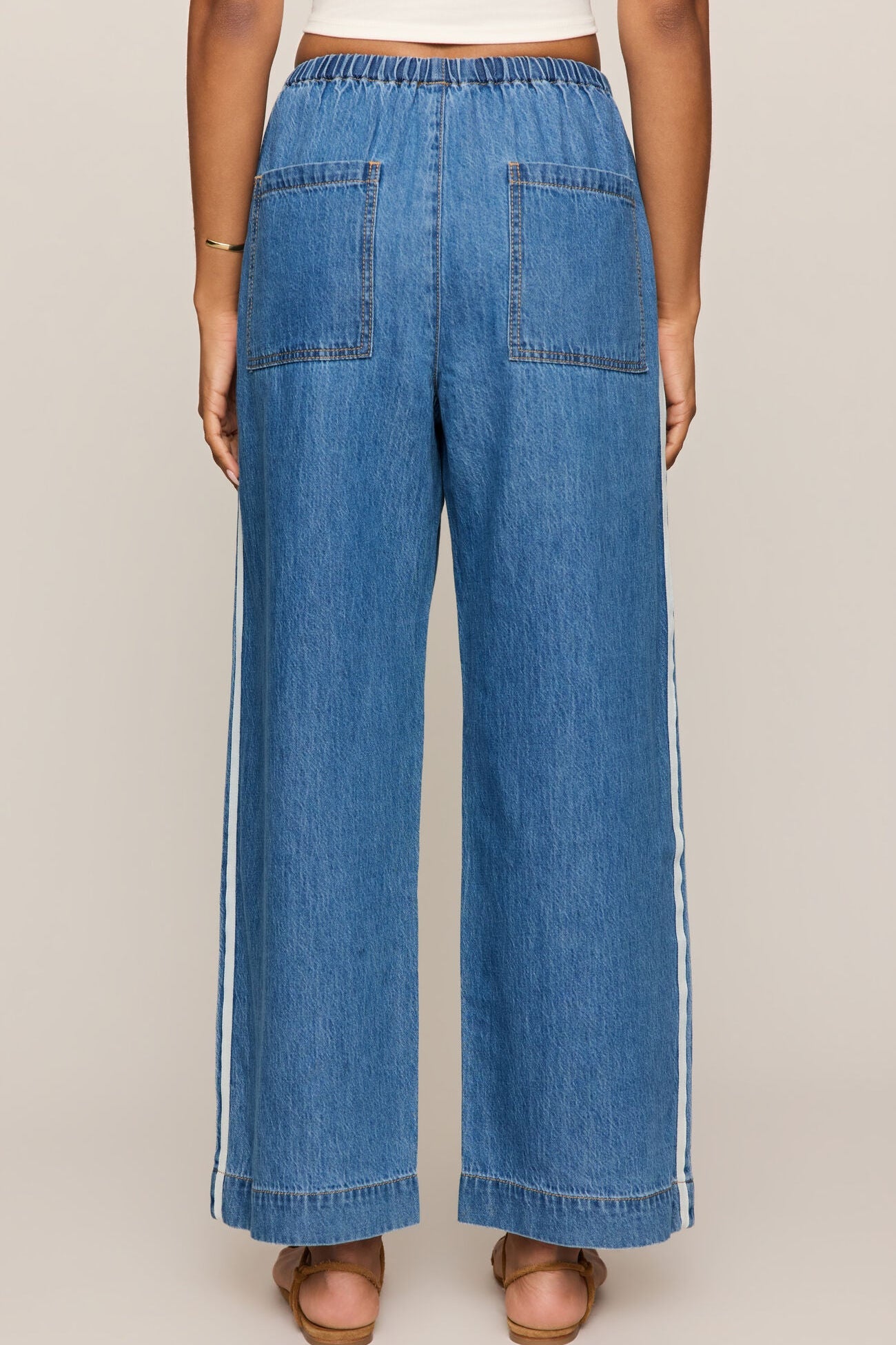 Back view of Woman wearing a white tank top and sidelines denim track pant on a beige background