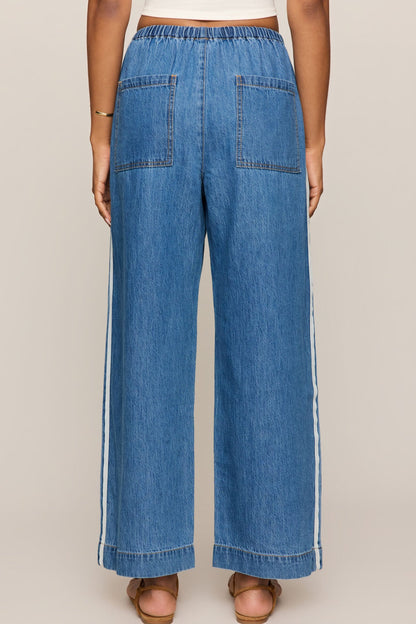 Back view of Woman wearing a white tank top and sidelines denim track pant on a beige background