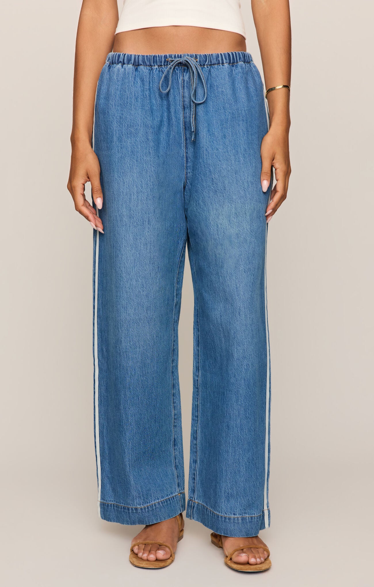 Front view of Woman wearing a white tank top and sidelines denim track pant on a beige background